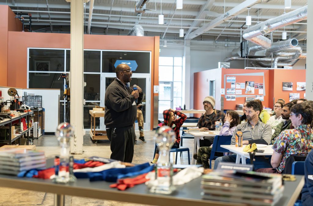 A man stands and speaks with a microphone to a seated audience in a modern, open workspace. In the foreground are trophies and ribbons on a table, capturing an awards or recognition moment for the demo day 2025 photo gallery.