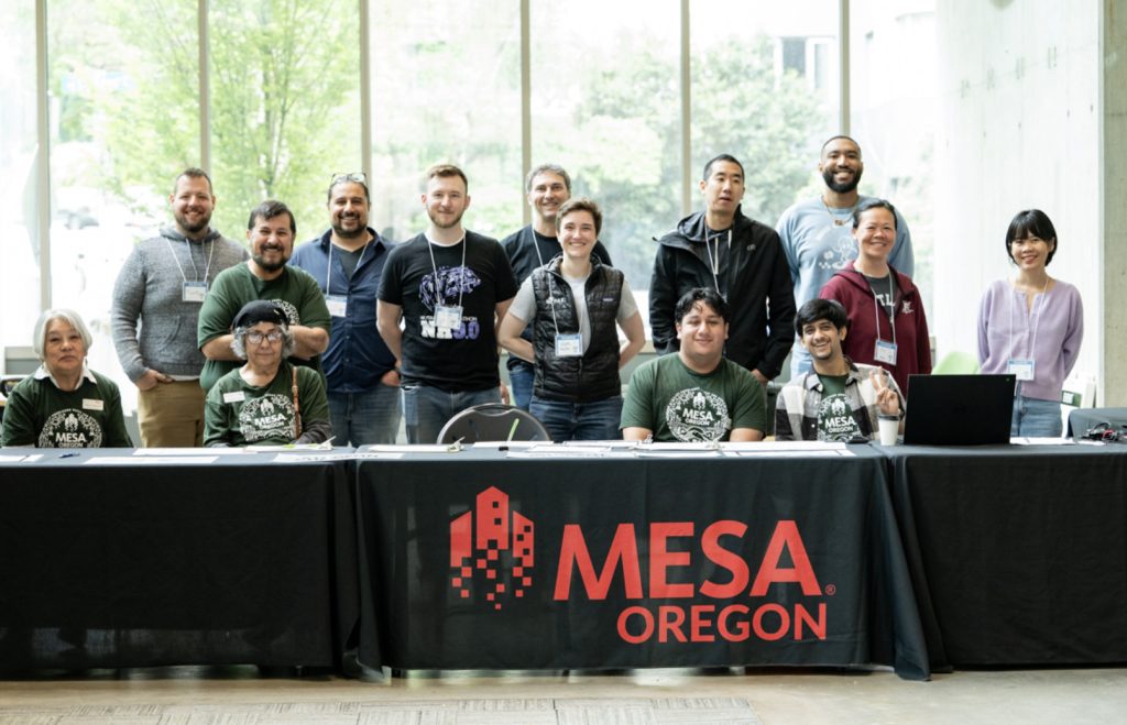 A group of people, some seated and some standing, smile behind a table with a black tablecloth displaying the MESA Oregon logo. Large windows in the background let in natural light at this Mesa Day 2025 photo gallery.