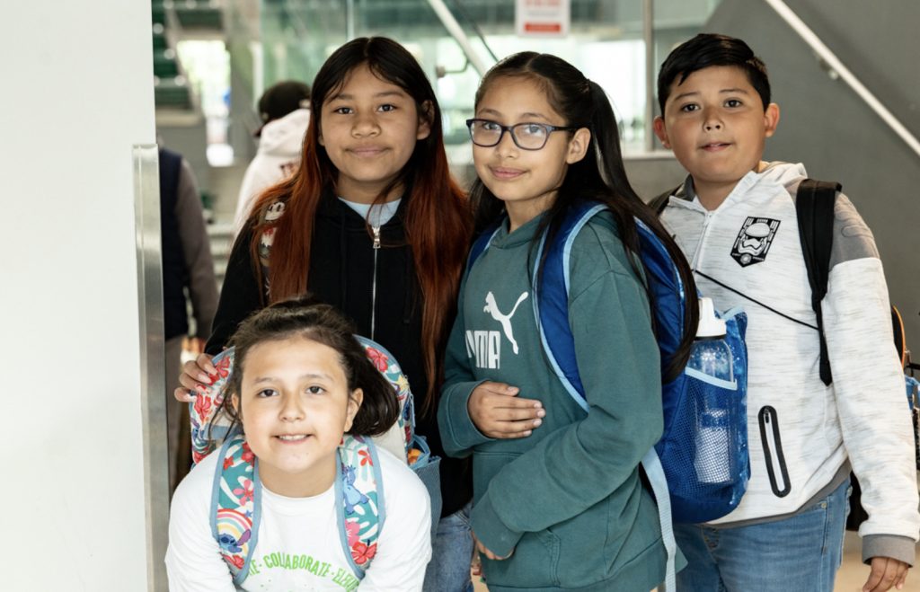 Four children stand indoors wearing backpacks and casual clothes, smiling for the camera. They appear to be in a school or public building with stairs and glass walls—perfect for the Mesa Day 2025 photo gallery.