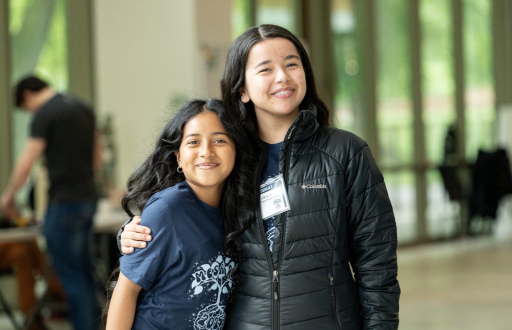 Two smiling girls stand together indoors, arm-in-arm and posing happily. One wears a navy t-shirt, the other a black jacket and name badge. The blurred background hints at large windows and people—captured for the Mesa Day 2025 photo gallery.