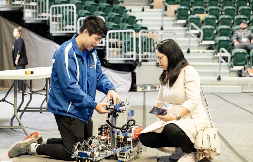 A young man and woman kneel on the ground in an arena, examining and adjusting a complex robot. The background shows empty green stadium seats and a few people sitting—an exciting moment captured for the mesa day 2025 photo gallery.