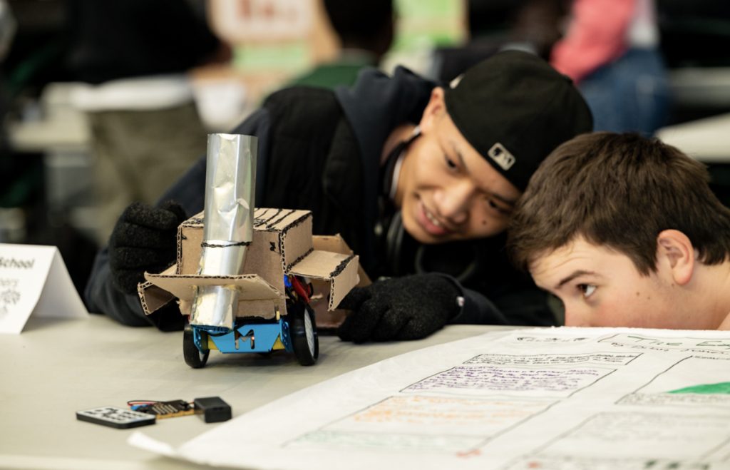 Two boys intently observe a small robot made of cardboard and metal parts on a table during the Mesa Day 2025 photo gallery, with one crouched at eye level while the other smiles and leans in, both fully engaged in a STEM activity.