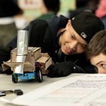 Two boys intently observe a small robot made of cardboard and metal parts on a table during the Mesa Day 2025 photo gallery, with one crouched at eye level while the other smiles and leans in, both fully engaged in a STEM activity.
