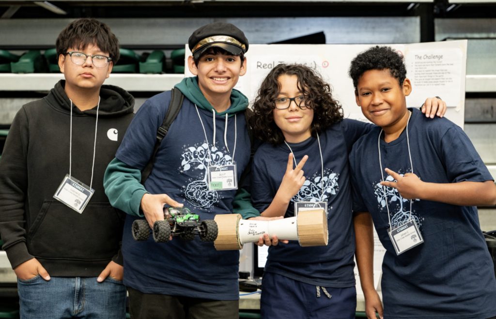 Four boys wearing name tags and matching blue T-shirts stand together in a gym. Two hold a small wheeled robot, and one makes a peace sign. They smile for the camera at Mesa Day 2025, featured in the photo gallery of this science event.