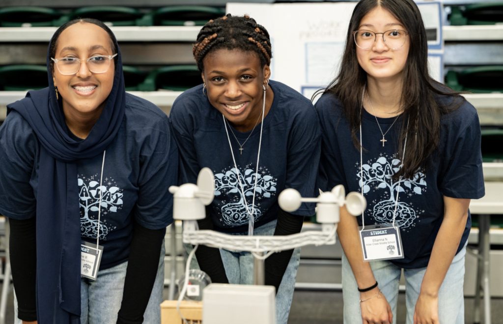 Three students wearing matching navy blue shirts and name tags smile while standing behind a science project display at the Mesa Day 2025 photo gallery, possibly during a science fair or exhibition.