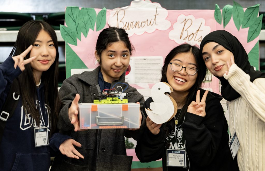 Four young women pose in front of a colorful Burnout Bots project display at the Mesa Day 2025 photo gallery; two make peace signs, one holds a small robot, and another holds a swan-shaped cutout. All are smiling.