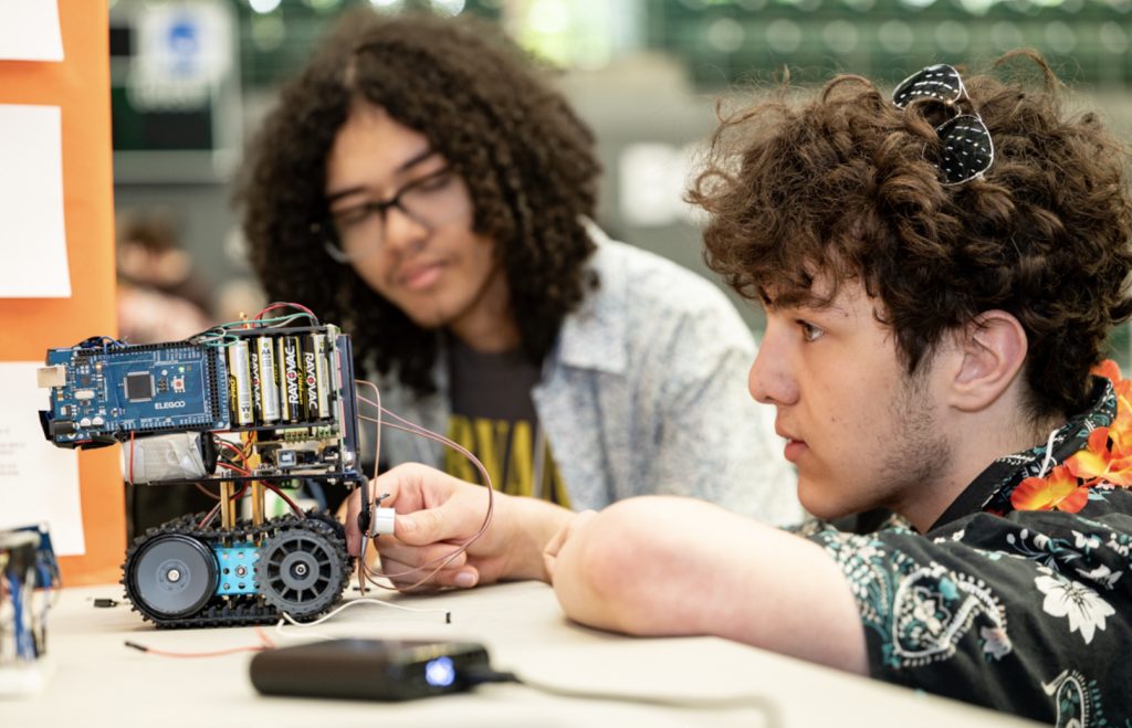 Two students work together on a small tracked robot at the table, adjusting its electronic components and wiring. This moment from the mesa day 2025 photo gallery shows one student observing while the other makes precise adjustments.