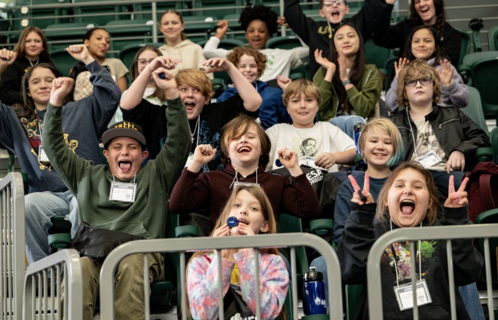 A group of enthusiastic children sit in stadium seats, cheering and smiling at the camera for the Mesa Day 2025 photo gallery. Some make peace signs, raise their arms, or hold up whistles, creating a lively and joyful atmosphere.