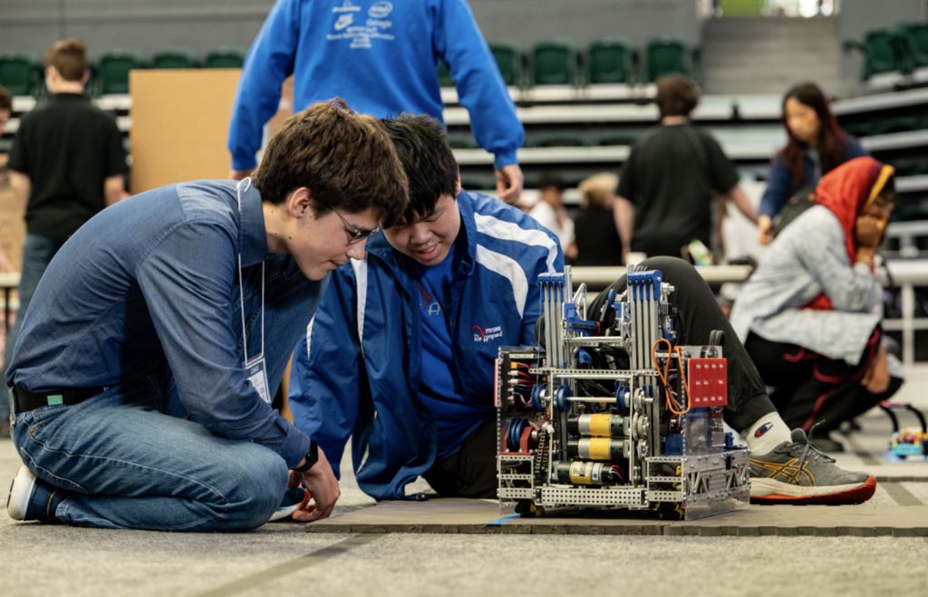 Two students kneel on the floor closely examining a complex robot during the Mesa Day 2025 competition. Other participants and robots can be seen in the background of the large indoor arena, captured for the event’s photo gallery.