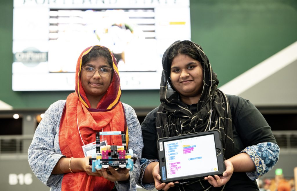 Two young women wearing hijabs stand indoors, smiling at the camera. One holds a colorful robotic device made of blocks, while the other displays a tablet with a coding program—captured for the mesa day 2025 photo gallery. A blurred event screen is behind them.