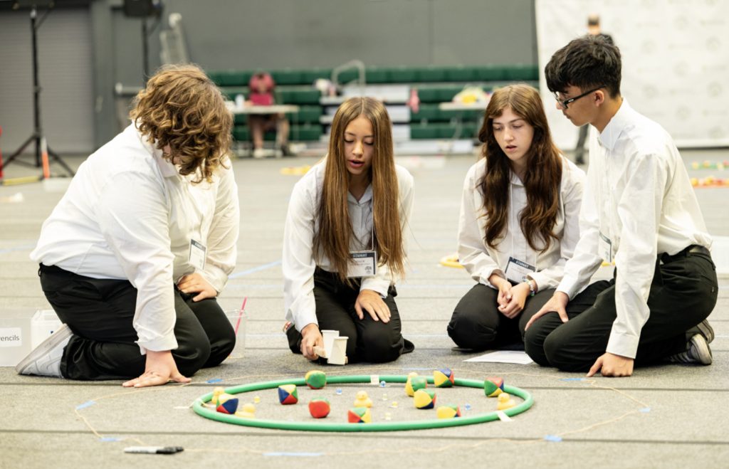 Four students in white shirts sit and kneel on the floor around a circular mat with colorful geometric objects, appearing to discuss or analyze them at Mesa Day 2025—captured for the event’s dynamic photo gallery.