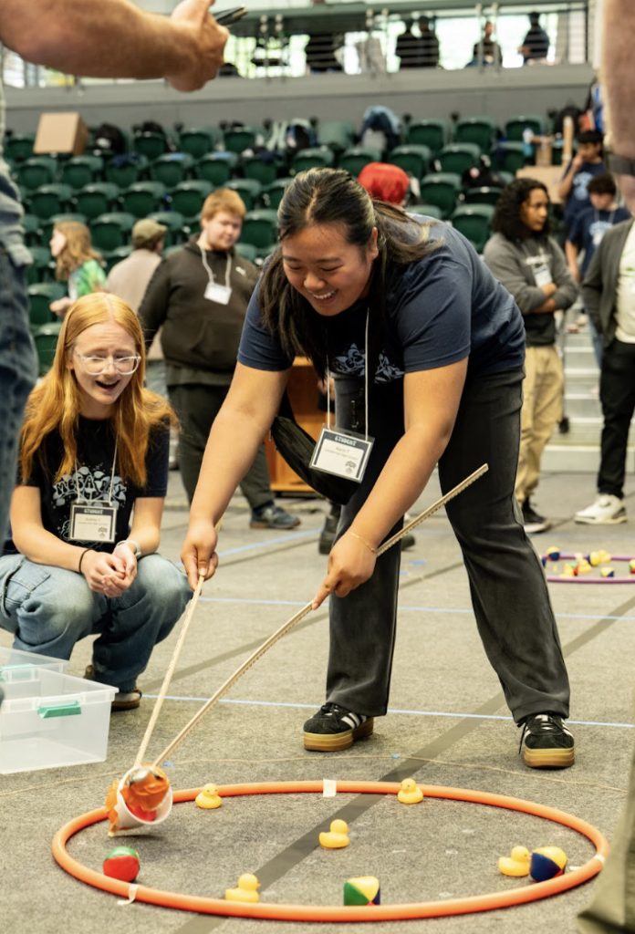 Two young people, one standing and one kneeling, use wooden sticks to move colorful objects inside a red hoop on the floor at a science or robotics competition—captured in this Mesa Day 2025 photo gallery, with other participants blurred in the background.