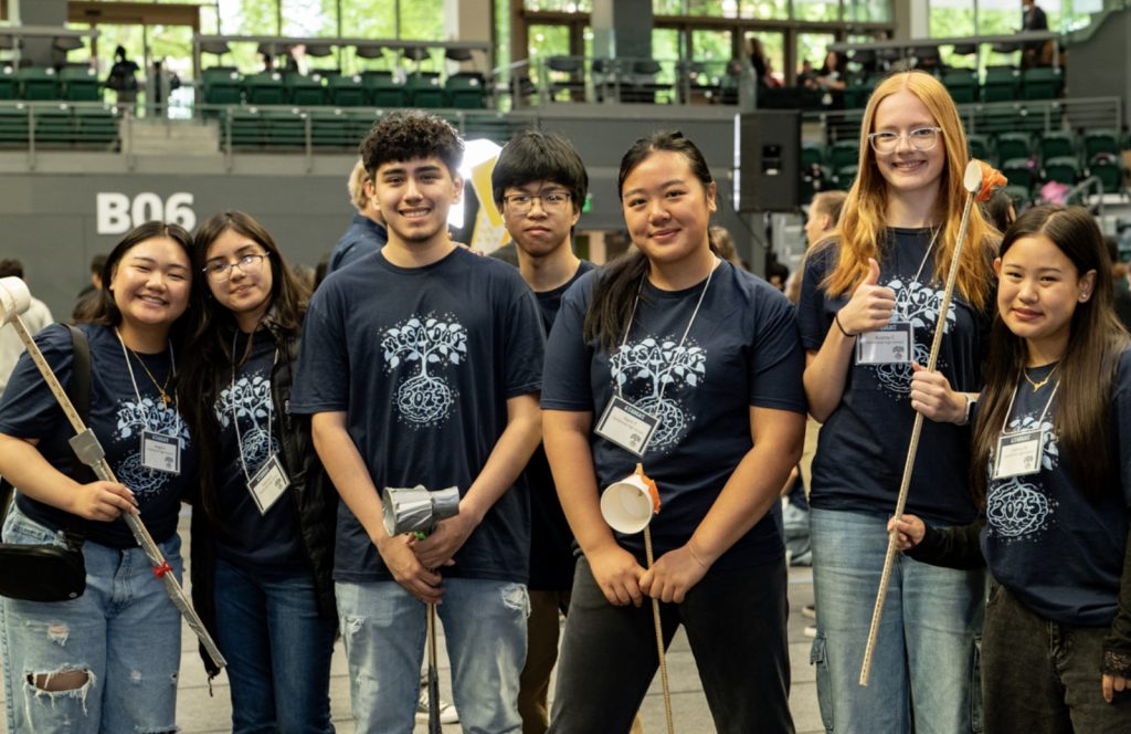 A group of seven smiling students wearing matching navy blue shirts and name badges stand indoors, holding handcrafted objects, with bleachers and other people in the background—captured in this Mesa Day 2025 photo gallery.