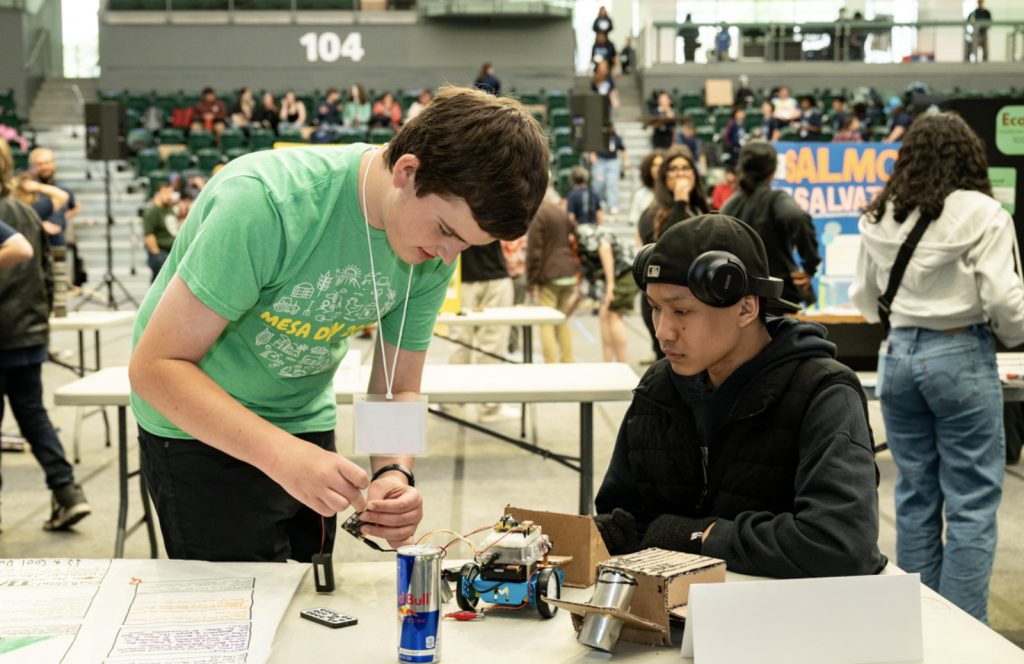 A student in a green shirt helps another in a black vest and headphones assemble a small robot at the busy gymnasium during Mesa Day 2025. Explore more moments like this in our Mesa Day 2025 photo gallery.