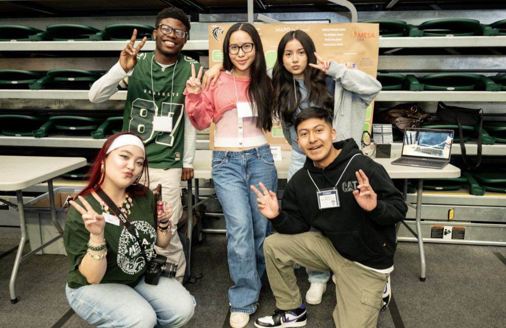 Five students pose and smile for the camera, flashing peace signs in front of their display booth with a laptop and informational poster. This mesa day 2025 photo gallery moment is set inside a gymnasium with empty green bleachers behind them.