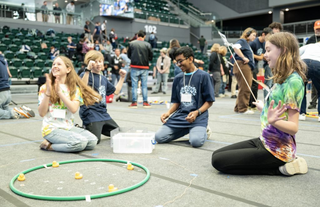 Four teenagers sit on a gym floor, smiling and playing a game with rubber ducks and a green ring as a crowd watches. The lively, friendly atmosphere is captured in this Mesa Day 2025 photo gallery moment.