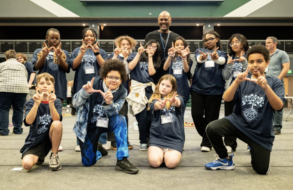 A group of diverse children and an adult stand indoors, smiling and posing with their hands making “W” shapes for the Mesa Day 2025 photo gallery. The children wear matching navy blue shirts and name tags, with some kneeling in front and others standing behind.