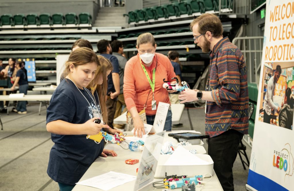 A girl examines a LEGO robotics creation at a booth while a man and woman assist her. Several people are in the background, and a "Welcome to LEGO Robotics" sign is visible on the right in this Mesa Day 2025 photo gallery.