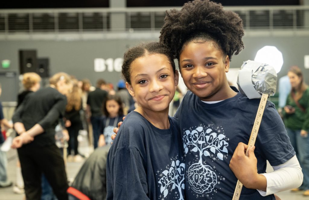 Two smiling girls in matching navy blue T-shirts pose together at an indoor event for the mesa day 2025 photo gallery; one holds a handmade hammer prop while people chat in the blurred background.