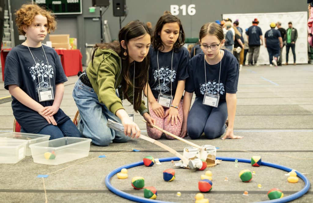Four young girls kneel on the floor at the MESA Day 2025 competition, guiding a handmade device to pick up objects inside a blue circle, surrounded by colorful balls and rubber ducks. All wear navy event T-shirts and name tags for the photo gallery.