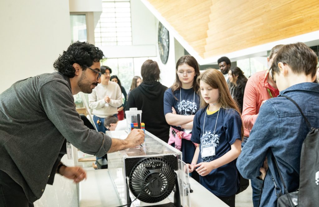 A man demonstrates a science experiment with a fan and clear container to a group of children and adults at an indoor event, as seen in the engaging mesa day 2025 photo gallery. The children watch attentively in the bright atmosphere.
