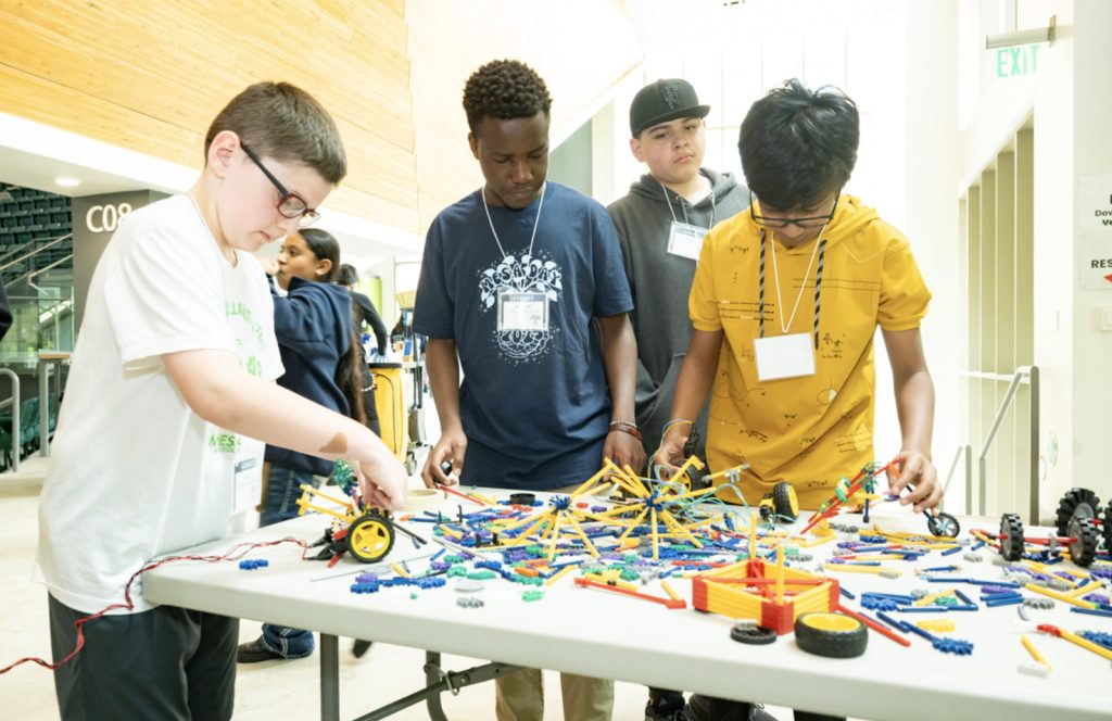 Four boys collaborate on a STEM project using colorful construction pieces at a table in a bright, modern building. Wearing name tags and casual clothes, they focus intently—perfect for the Mesa Day 2025 photo gallery.