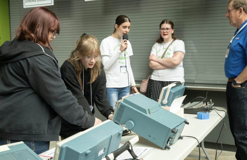 A group of young people and an adult stand around a table with vintage electronic equipment at the Mesa Day 2025 photo gallery. One girl speaks into a microphone while others look at the devices, engaged in a hands-on activity or demonstration.