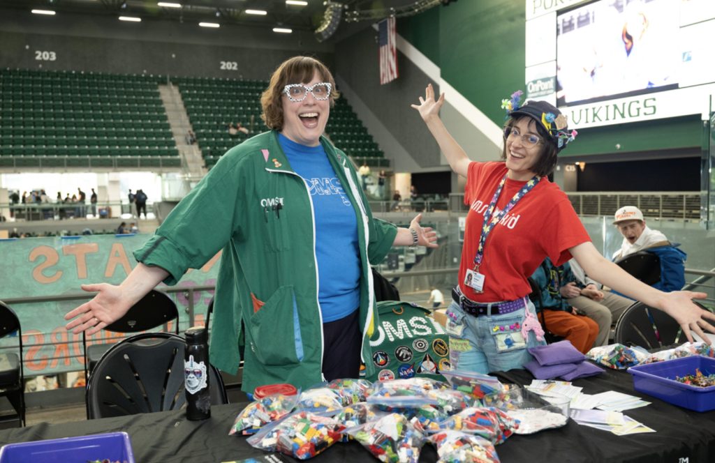 Two smiling women stand behind a table filled with colorful LEGO pieces at Mesa Day 2025, arms open wide. One wears a green jacket with patches, the other a red shirt and decorated hat. Bleachers are visible behind them in this photo gallery moment.