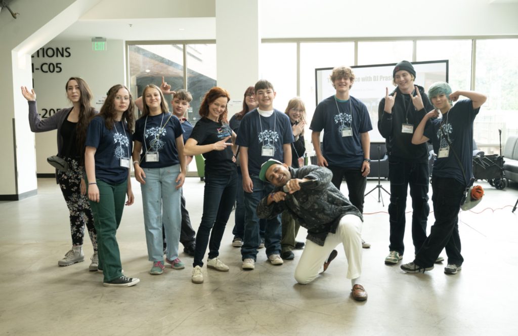 A group of teens and adults wearing matching navy t-shirts pose playfully indoors for the Mesa Day 2025 photo gallery, some making hand signs and funny faces as bright natural light streams through large windows behind them.