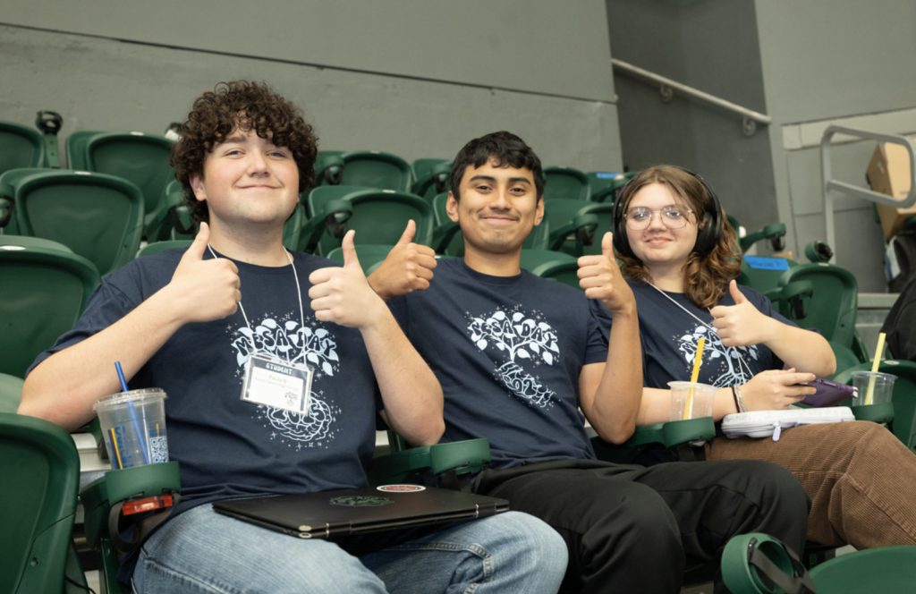 Three young people sit in green stadium seats, smiling and giving thumbs up at Mesa Day 2025. They wear matching navy blue shirts with white designs and name tags. Two have drinks; one has a laptop. Indoors, casual, cheerful photo gallery vibe.