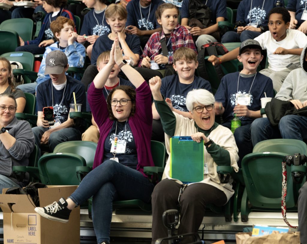 A group of people, including excited children and cheering adults, sit in green arena seats at a Mesa Day 2025 event. Two women in the front row celebrate enthusiastically, while others smile and watch the action for the photo gallery.