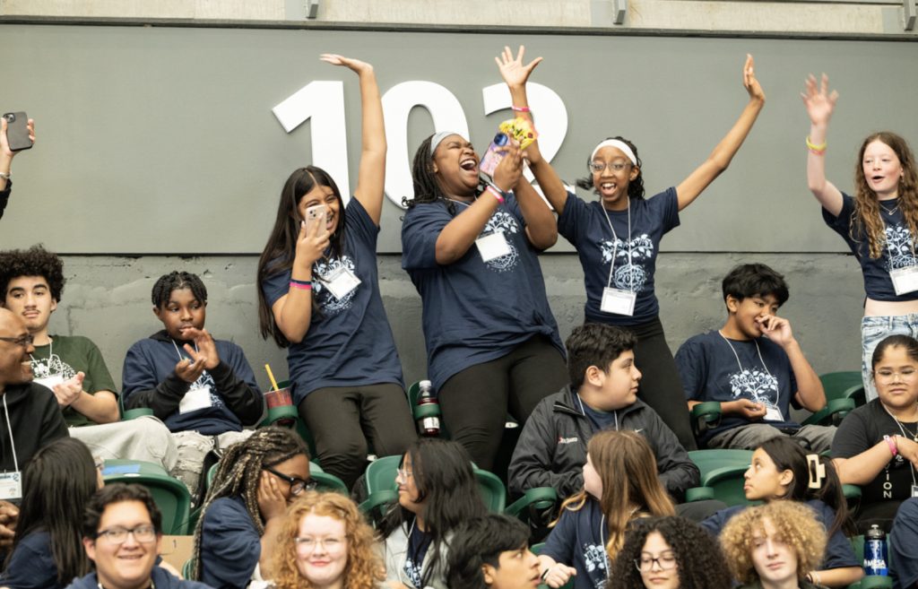 A diverse group of excited students in matching blue shirts cheer and celebrate in stadium seats at Mesa Day 2025, with two girls raising their arms while others smile, laugh, or take photos for the event’s photo gallery.