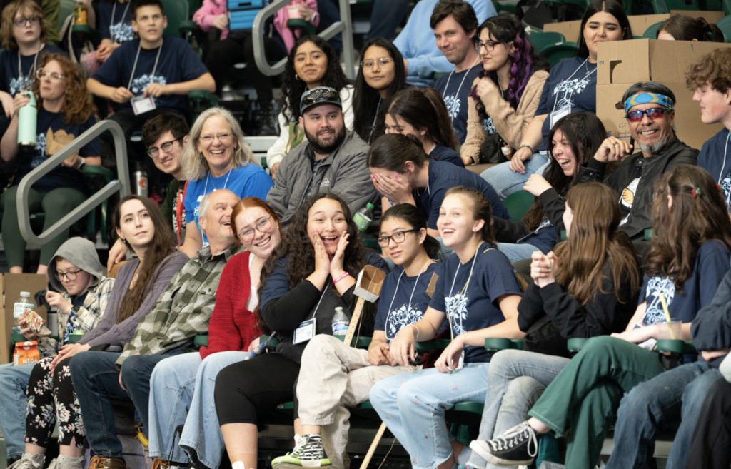 A large group of people, mostly young adults and teens, sit together in stadium seats, smiling and interacting. Many wear matching navy blue shirts and event badges, capturing the lively spirit featured in the Mesa Day 2025 photo gallery.