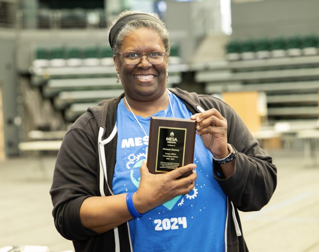 A smiling woman wearing glasses and a blue MESA 2024 shirt holds a black and gold plaque award in a large indoor venue with empty bleachers, perfect for the mesa day 2025 photo gallery.