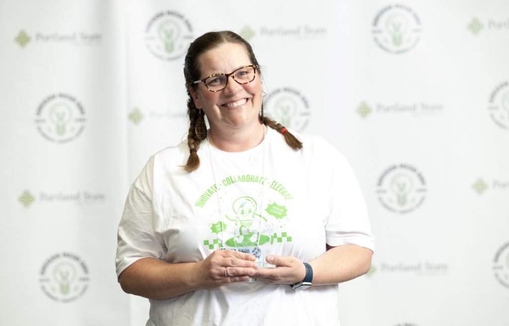 A smiling woman with glasses and braids holds an award at Mesa Day 2025. She wears a white T-shirt with a green logo and stands in front of a Portland State University logo backdrop for the photo gallery.