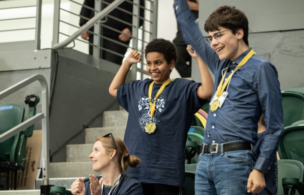 Two boys wearing medals celebrate with raised arms beside a smiling woman seated in front of them at the indoor arena, with green seats and a staircase behind—in this joyful Mesa Day 2025 photo gallery moment.
