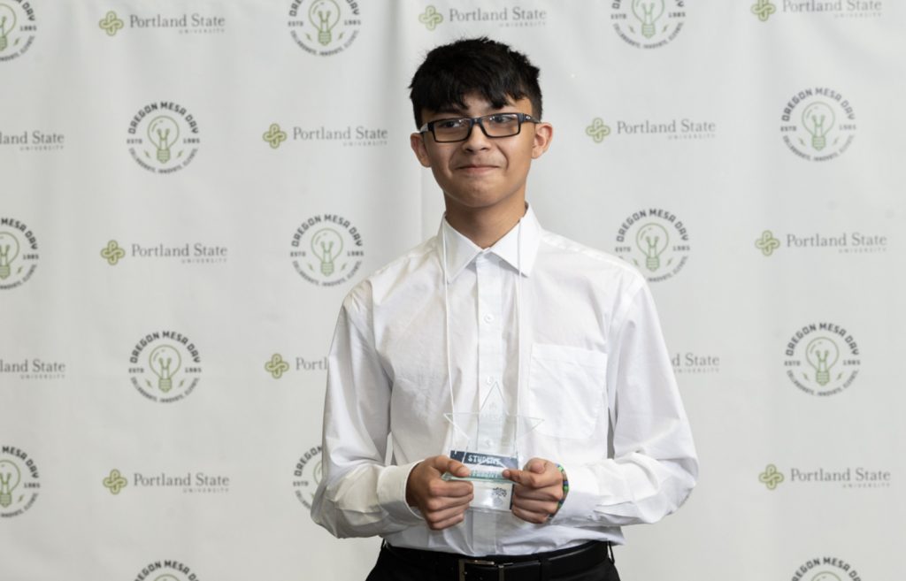 A young person wearing glasses and a white shirt stands in front of an Oregon MESA and Portland State University backdrop, holding a clear award and smiling slightly at the camera—captured for the Mesa Day 2025 photo gallery.