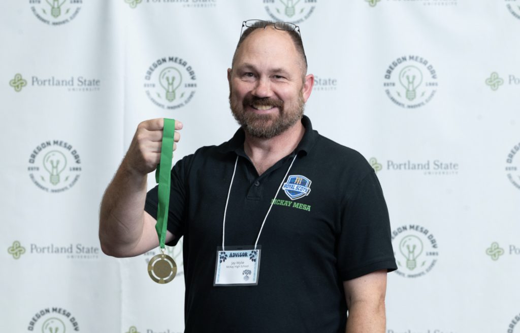 A man with a beard, wearing a black polo shirt and name tag, smiles while holding up a gold medal. The background features Portland State University and Oregon MESA Day logos from the Mesa Day 2025 photo gallery.