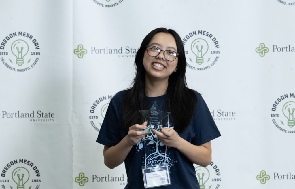 A smiling young woman holds a glass award in front of an Oregon Mesa Day and Portland State University backdrop. She wears glasses, a dark blue T-shirt, and a name badge—perfect for the Mesa Day 2025 photo gallery.