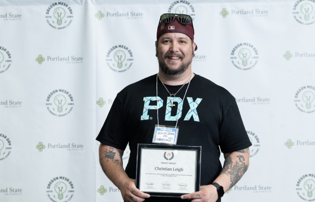 A smiling man in a red cap and PDX t-shirt holds a certificate, standing before a backdrop with Portland State University and Oregon MESA Day logos—perfect for the Mesa Day 2025 photo gallery.