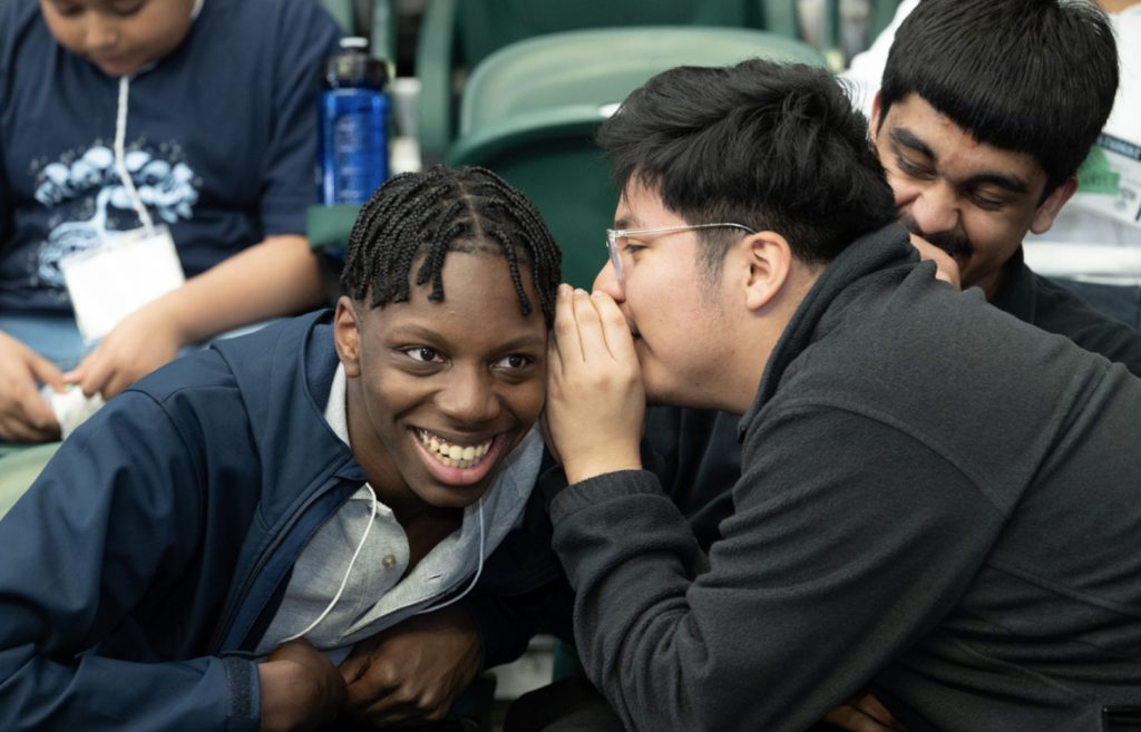 A group of students sit together. One whispers into another’s ear while both smile and laugh, and a third student behind them also laughs, capturing a friendly, lighthearted moment at the mesa day 2025 photo gallery.