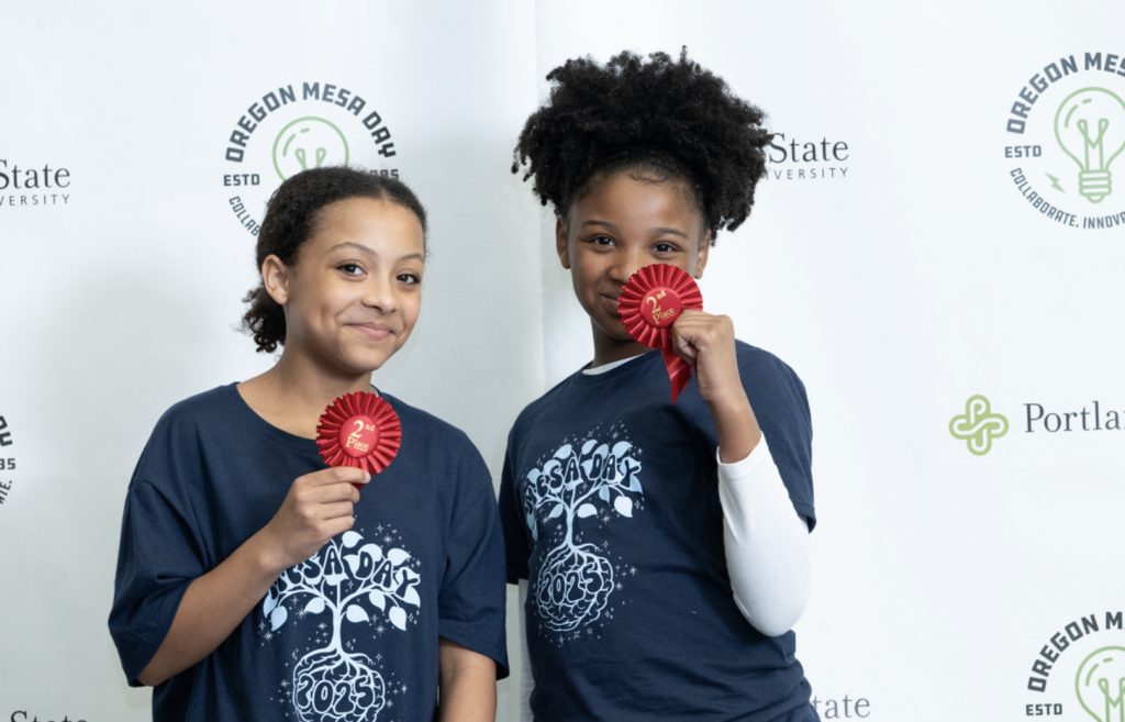 Two young girls smiling and holding red award ribbons in matching navy Oregon MESA Day t-shirts, standing before a backdrop with Oregon MESA and sponsor logos—a joyful moment captured for the Mesa Day 2025 photo gallery.