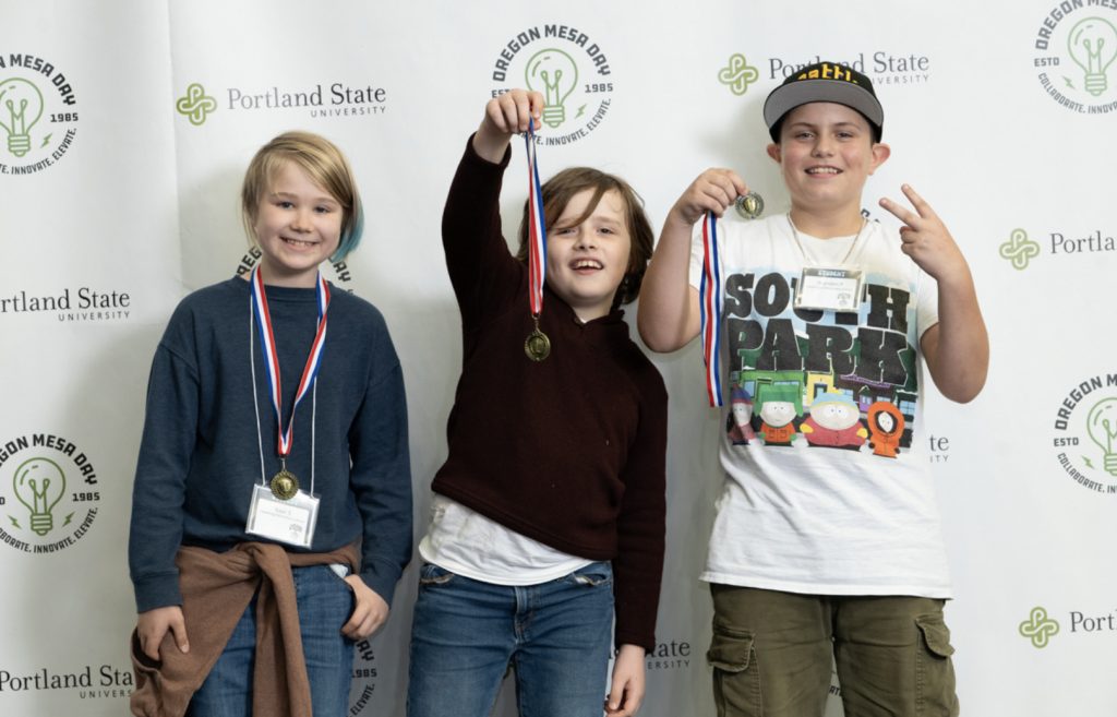 Three children smiling and posing with medals in front of a Portland State University and Oregon MESA Day backdrop; one holds up a medal, another flashes a peace sign. Featured in the MESA Day 2025 photo gallery, all wear name tags.