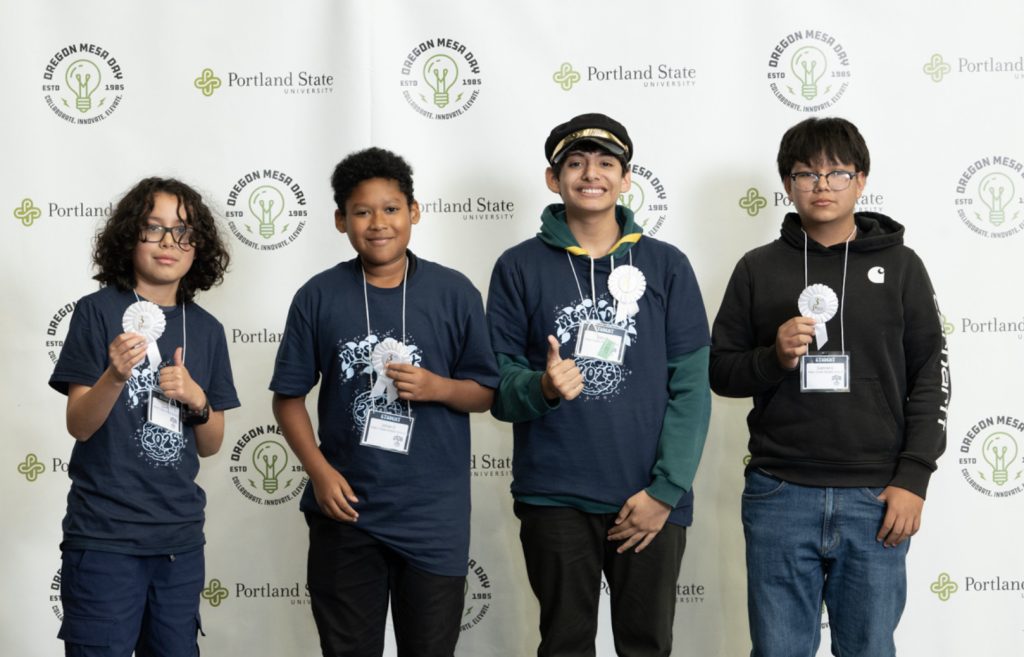 Four boys pose in front of a Portland State University and Oregon MESA backdrop, smiling and holding medals—three in navy blue event t-shirts, one in a black hoodie—for the Mesa Day 2025 photo gallery.