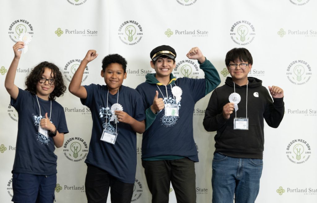 Four smiling boys wearing medals and navy blue t-shirts stand in front of a white backdrop with Portland State and Oregon MESA Day logos, raising their hands in celebration for the Mesa Day 2025 photo gallery.
