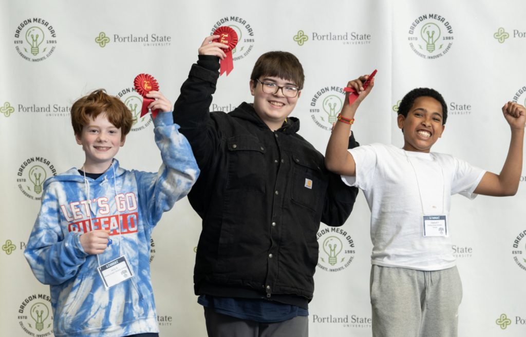 Three smiling boys stand in front of a Portland State University and Oregon MESA backdrop, holding red ribbons and wearing name tags, celebrating their achievement at the Mesa Day 2025 photo gallery.