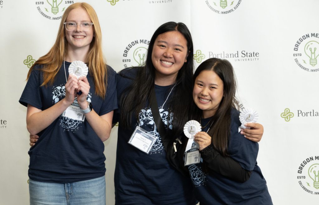 Three smiling young women in navy blue shirts and name tags hold white award ribbons, standing before an Oregon State and Portland State University backdrop at the Mesa Day 2025 photo gallery.