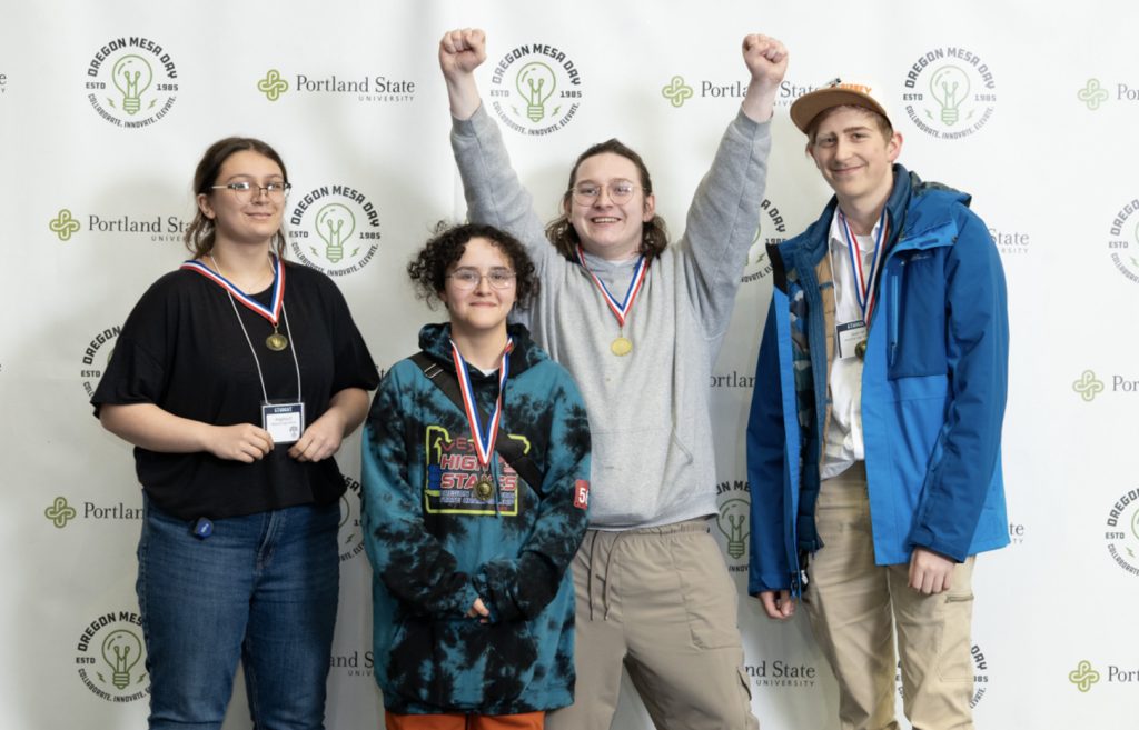 Four people wearing medals stand in front of a Portland State University backdrop; one person raises both arms in celebration while the others smile at the camera in this Mesa Day 2025 photo gallery.