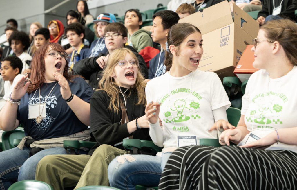 A group of enthusiastic students sit in stadium seats, smiling and cheering. The front row features four girls, two in white MESA shirts, reacting excitedly at Mesa Day 2025. In the background, more students watch the event—perfect for a photo gallery.