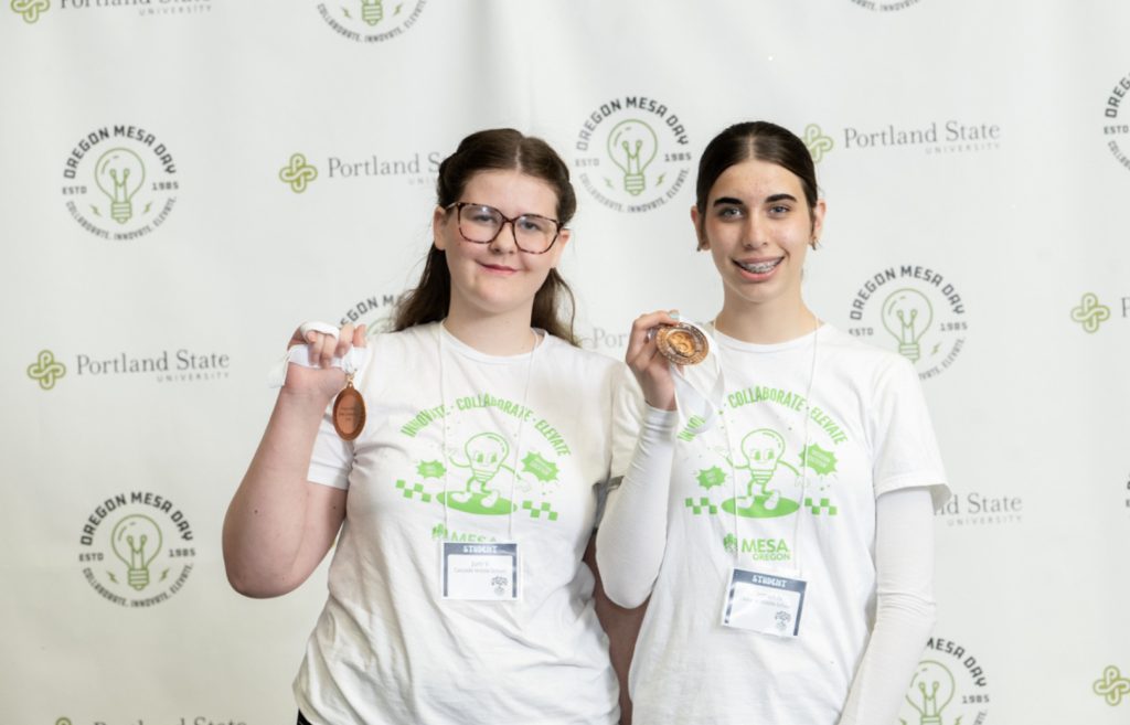 Two young women wearing glasses and white Oregon MESA t-shirts stand in front of a step-and-repeat banner, smiling and holding up bronze medals. Both have name badges and look proud—captured for the Mesa Day 2025 photo gallery.